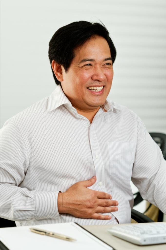 Ahmad, a middle-aged Asian man, smiling and looking relaxed while sitting comfortably at his desk, gesturing towards his lower abdomen area.
