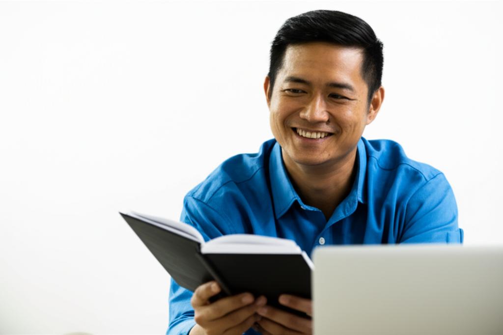 Prompt: A middle-aged Malaysian man smiling and looking comfortable while sitting, perhaps enjoying a hobby like reading or using a laptop, bright and relaxed atmosphere.